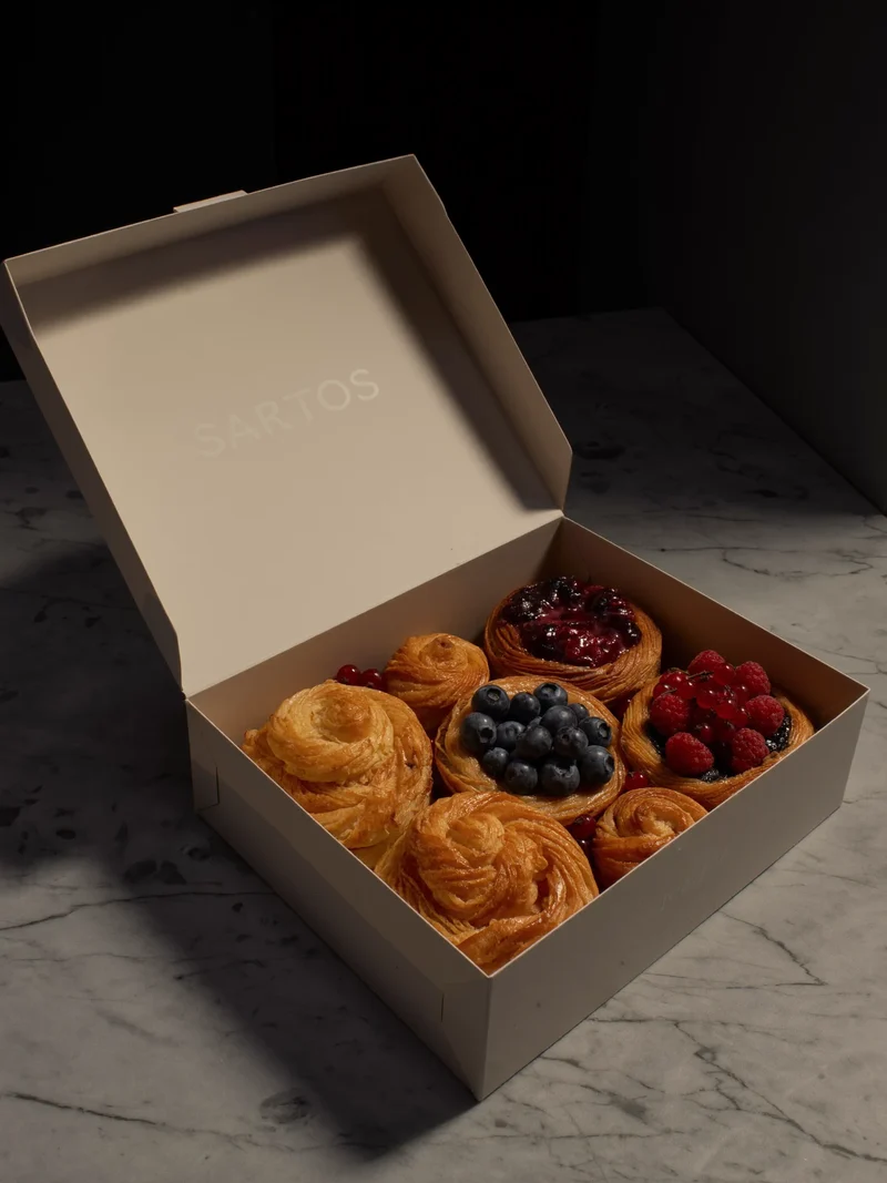 Kleine Gebäck- und Beeren-Tartelette-Auswahl in neutraler Dessertbox auf hellem Marmor